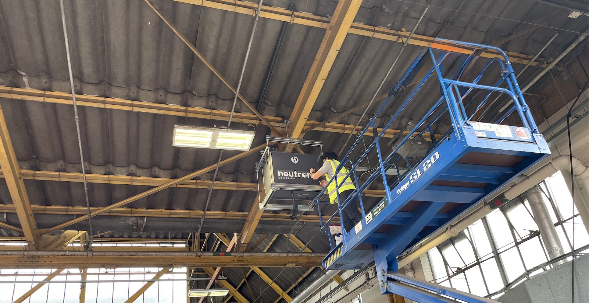Neutron Systems engineer on a scissor lift installing a Neutron NSNF0014DDC overhead drop-down connector unit to the structural steelwork of a covered bus depot